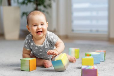 Puantiyeli neşeli bir bebek yerde oturuyor ve parlak oturma odasında renkli yumuşak küplerle oynuyor..
