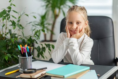 Beyaz bluzlu mutlu genç kız masasında gülümsüyor, parlak bir odada renkli kalemler, defterler ve eğitici aletlerle çevrili..