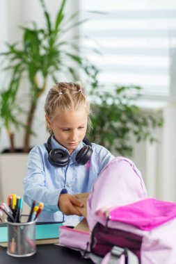 Kulaklıklı genç bir öğrenci çalışma masasında okula hazırlanırken not defterlerini pembe bir sırt çantasına dikkatlice yerleştirir..