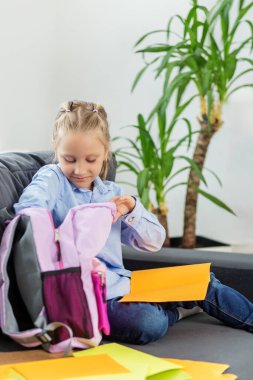 Genç bir kız kanepeye oturur parlak kağıt klasörlerini düzenler ve onları açık pembe sırt çantasına yerleştirir..