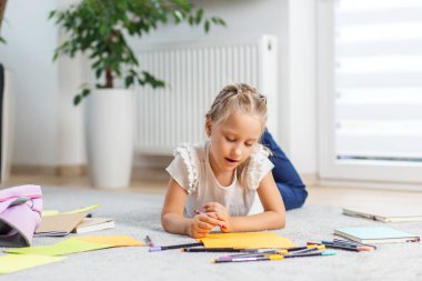 Genç kız halıya uzanmış, parlak turuncu kağıtlara kalemler çizmeye odaklanmış, etrafı kitaplar ve okul malzemeleriyle çevrili..
