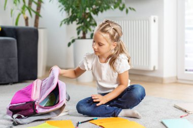 Genç kız halı üzerinde bağdaş kurarak oturuyor, parlak bir ev ortamında açık sırt çantasına not defterleri ve renkli dosyalar yerleştiriyor..