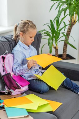 Genç kız elinde parlak kağıt klasörlerle kanepede oturur ve evdeki açık pembe sırt çantasının yanında okul malzemelerini düzenler..