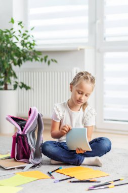 Genç bir kız, elinde mavi bir defterle bir halının üzerinde oturuyor. Etrafı okul gereçleri ve evde açık bir sırt çantasıyla çevrili..