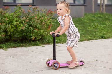 Çizgili kıyafetli genç çocuk, yerleşim alanındaki yeşil bir yolda üç tekerlekli pembe bir scooter sürmekten hoşlanıyor..