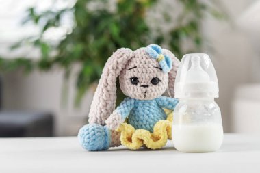 Mavi elbiseli yumuşak el yapımı doldurulmuş tavşan bulanık arka planı olan beyaz bir yüzeyde süt dolu bir bebek şişesinin yanında oturuyor..