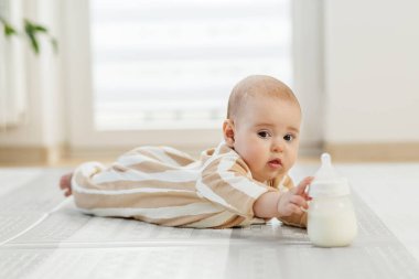Çizgili pijamalı bir bebek, sütle dolu bir bebek beslenme şişesine dokunmak için yere uzanır..