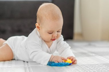 Beyaz elbiseli bir bebek yerde yatıyor, ilginç bir şekilde mavi ve sarı bir diş çıkarma oyuncağıyla oynuyor..