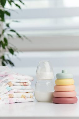 Düzenli katlanmış bebek kıyafetleri ve renkli istifleme yüzüklerinin yanına yerleştirilmiş sütle dolu bir bebek besleme şişesi, çocuk bakımını ve ebeveynliği sembolize ediyor..
