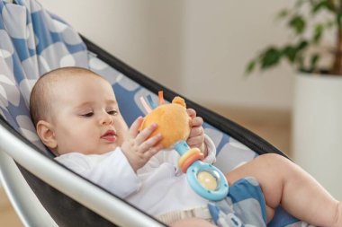 Yastıklı bir fedaide oturan sevimli bebek, doğal ışıkta dinlenirken renkli bir çıngırak oyuncağını tutuyor ve keşfediyor..