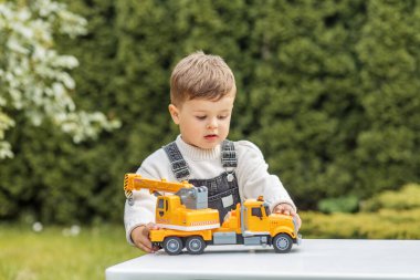 Tulum giymiş genç bir çocuk açık bir masada parlak sarı oyuncak bir inşaat kamyonu ile oynuyor, yaratıcı oyun zamanlarının keyfini çıkarıyor..