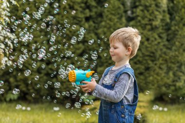 Gülümseyen genç çocuk bahçede güneşli bir günün tadını çıkarıyor, elinde yüzen sabun köpükleriyle çevrili bir baloncuk tabancası tutuyor..