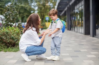 Gülümseyen bir anne, okula gitmeden önce mutlu bir anı paylaşarak, neşeli oğlunun önünde çömelir..