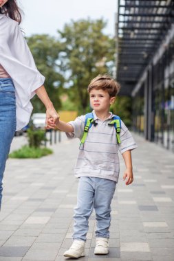Sırt çantalı genç bir çocuk annesinin elini tutarak şehir kaldırımında yürüyor ve kararlılıkla okula gidiyor.
