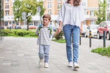Sırt çantalı genç bir çocuk güneşli bir günde okula giderken annesiyle kaldırımda el ele yürür..