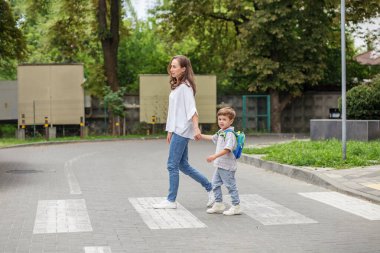 Sırt çantalı bir anne ve küçük oğlu, bir şehir caddesinde yaya geçidinde el ele yürüyorlar..