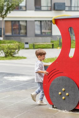 Günlük kıyafetlerle gezen küçük bir çocuk, tren şeklinde kırmızı bir oyun alanını keşfediyor, apartmanın yakınında dışarıda oyun oynamaktan zevk alıyor..