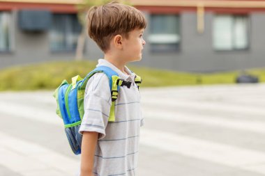 Çizgili tişört ve sırt çantası giymiş, güneşli bir günde okul binasının yanında duran genç bir çocuğun yan görüntüsü..