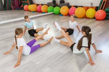 Bir grup genç kız daire şeklinde bacak kaldırma egzersizleri yapıyor, eğlenceli bir kapalı spor salonunda güç ve koordinasyon üzerine çalışıyorlar..