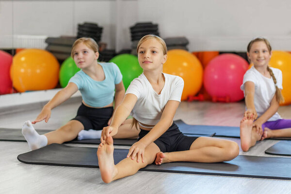 Три молодые девушки выполняют сидячие растяжки для ног во время занятий йогой или гибкостью, улучшая осанку и подвижность в помещении спортзала.