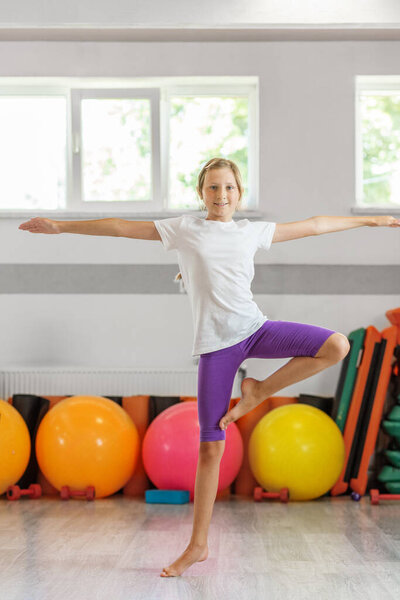 Уверенная девушка, держащая равновесие, позирует в тренажерном зале, окруженная тренажерами и яркими мячами для упражнений на заднем плане.