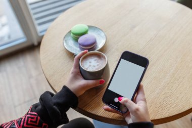 Tabağında renkli badem kurabiyeleri olan ahşap bir kafe masasında elinde bir fincan kahve ve akıllı telefon tutan birinin yakın çekimi..