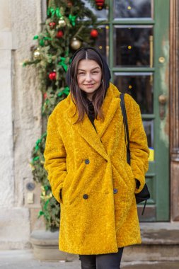 Tüylü sarı kışlık ceketli, kendine güveni tam bir kadın Noel süslerinin yanında poz veriyor ve klasik yeşil bir kapısı var..