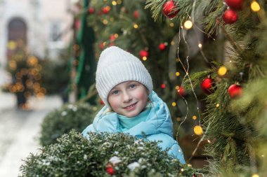 Açık mavi ceketli ve beyaz örgü şapkalı mutlu genç kız Noel ağaçları arasında kırmızı süsler ve ışıklarla poz veriyor..