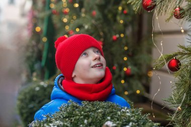 Kırmızı şapkalı ve mavi kışlık ceketli neşeli genç adam süslenmiş bir sokakta Noel süsleri ve ışıklara hayran..