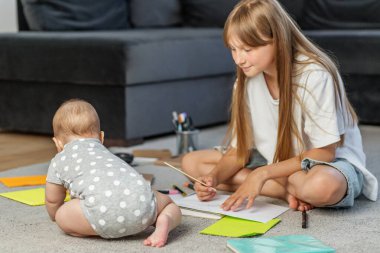 Genç bir kız halı üzerinde bacak bacak üstüne atmış, kağıt üzerinde resim çizerken, bebeği puantiyeli bebek tulumunun içinde etrafı keşfediyor..