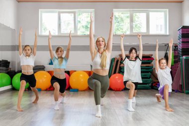 Kadın eğitmen, bir grup genç kıza yoga pozu veriyor. Kapalı bir stüdyoda renkli spor aletleri ve paspaslarla..
