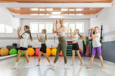 Çocuklar rengarenk bir spor salonunda senkronize el hareketleri yaparak dans eden bir hocayı takip ederler..