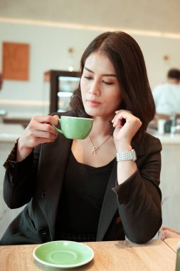 Siyah ceket giyen güzel bir kariyer kadını, bir kafede. Ellerinden biri yeşil bir kahve bardağı tutuyor..