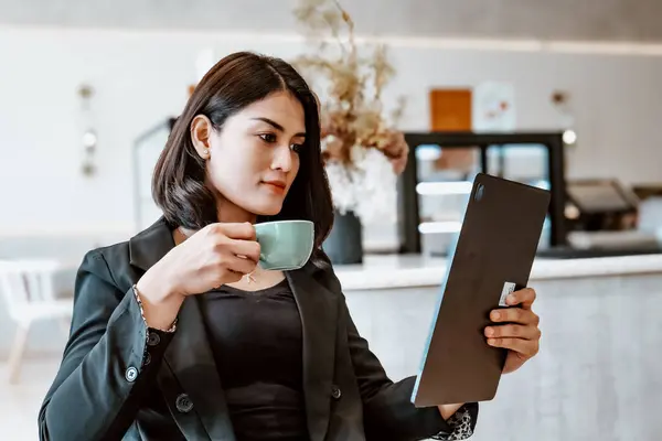 Siyah ceket giyen güzel bir kariyer kadını, bir kafede. Bir elinde yeşil bir kahve fincanı, diğerinde de bir tablet var..