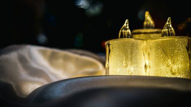 Arka planda parıldayan Noel ışıkları olan beyaz saten bir kumaşın üzerinde parlayan üç Noel mumu. Sol tarafta boş bir alan var, metin ile doldurulabilir..