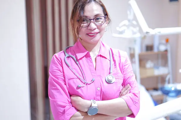 Pembe gömlekli bir tıp uzmanı klinik ortamında steteskop tutuyor. Hasta muayenesi için hazır. Arkaplanda tıbbi ekipmanlar yer alıyor, sağlık ortamının vurgulanması.