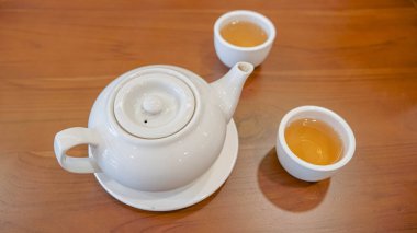 Uyumlu kapağı ve sapı olan beyaz seramik bir çaydanlık, açık kahverengi çayla dolu iki küçük beyaz fincanla birlikte ahşap bir masada oturur. Kurulum, sakin bir çay içme deneyimi öneriyor..