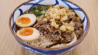 Bol haşlanmış yumurta, dilimlenmiş et, tempura taneleri, yeşil soğan ve susam tohumlarıyla kaplı bir kase Japon eriştesi canlı renkler ve dokular sergiliyor..