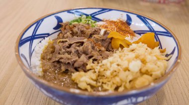 İçinde taze et dilimleri, beyaz pirinç, tempura parçaları, turşu zencefil, yeşil soğan ve geleneksel mavi bir kasede servis edilen bir tutam kırmızı biber tozu bulunan lezzetli bir Japon gyudonu..