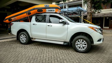 Yeşil ve diğer araçlarla çevrili bir otoparkta turuncu şişme bot taşıyan beyaz bir kamyonet. Maceraya hazırım..