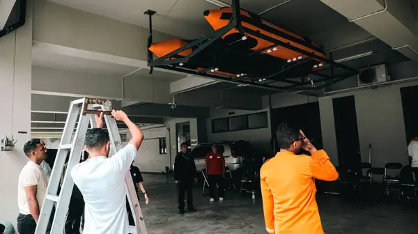 Merdivendeki bir adam, otoparkta turuncu şişme bir tekne tutan tavana monte edilmiş bir askıyı ayarlıyor..