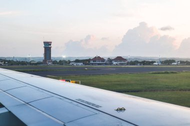 Görüntü bir uçak kanadı, kontrol kulesi ve gün batımında Bali 'deki Ngurah Rai Havalimanı' ndaki terminal binalarını gösteriyor..