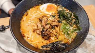 Bol haşlanmış yumurta, dilimlenmiş et, tempura taneleri, yeşil soğan ve susam tohumlarıyla kaplı bir kase Japon eriştesi canlı renkler ve dokular sergiliyor..