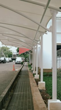 Görüntü, beyaz kolonları olan kapalı bir yürüyüş yolunu gösteriyor. Arabaları ve trafik konisi olan bir park alanına bitişik..