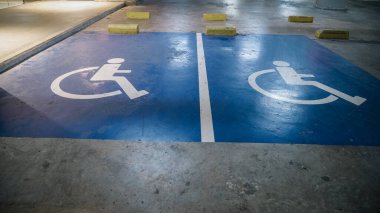 Bu görüntü, bir yeraltı otoparkında iki mavi engelli park alanını gösteriyor. Beyaz tekerlekli sandalye sembolleri ve sarı tekerlek duraklarıyla işaretlenmiş..