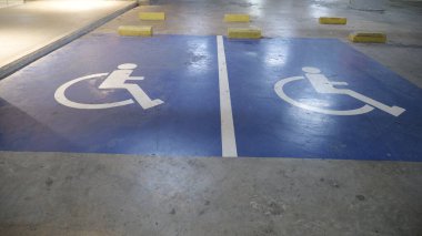 Bu görüntü, bir yeraltı otoparkında iki mavi engelli park alanını gösteriyor. Beyaz tekerlekli sandalye sembolleri ve sarı tekerlek duraklarıyla işaretlenmiş..