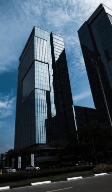 Uzun cam gökdelenler mavi gökyüzünü yansıtıyor, önplanda arabalar ve yeşillikler olan bir iş bölgesinde yer alıyor..