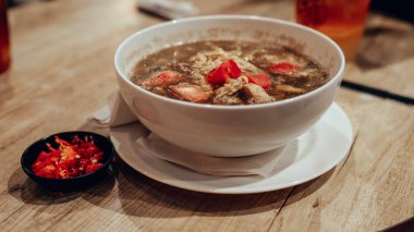 Buharda pişen bir çorba kasesine yakın plan et parçaları ve kırmızı biber, yanında küçük bir tabak doğranmış kırmızı biber, ahşap bir masaya dizilmiş..
