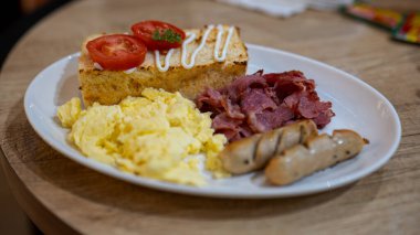 Çırpılmış yumurta, kıtır pastırma, ızgara sosis, üstüne domates ve mayonez konmuş beyaz bir tabakta servis edilen doyurucu bir kahvaltı..