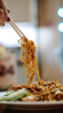 Yemek çubuklarının yakın çekimi sebze ve tavukla kızartılmış erişte kaldırma, salatalık dilimleri ile süslenmiş, lezzetli bir Asya mutfağı sergileniyor..